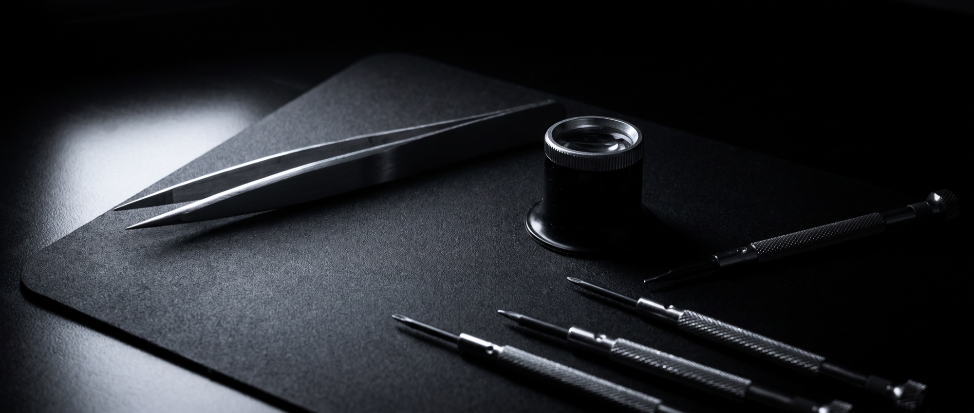 A watchmaker's bench in near darkness: steel tweezers and a loupe on a matte surface under a single cold light.